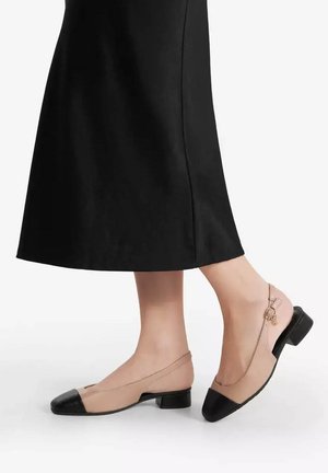 Pieds portant des chaussures plates beige et noires avec des brides à la cheville, associées à une jupe midi noire sur fond blanc.