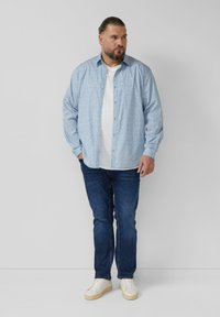 Lichtblauw overhemd met knopen en een klein bloemenpatroon, gedragen over een wit T-shirt, gecombineerd met donkere spijkerbroeken en witte sneakers.