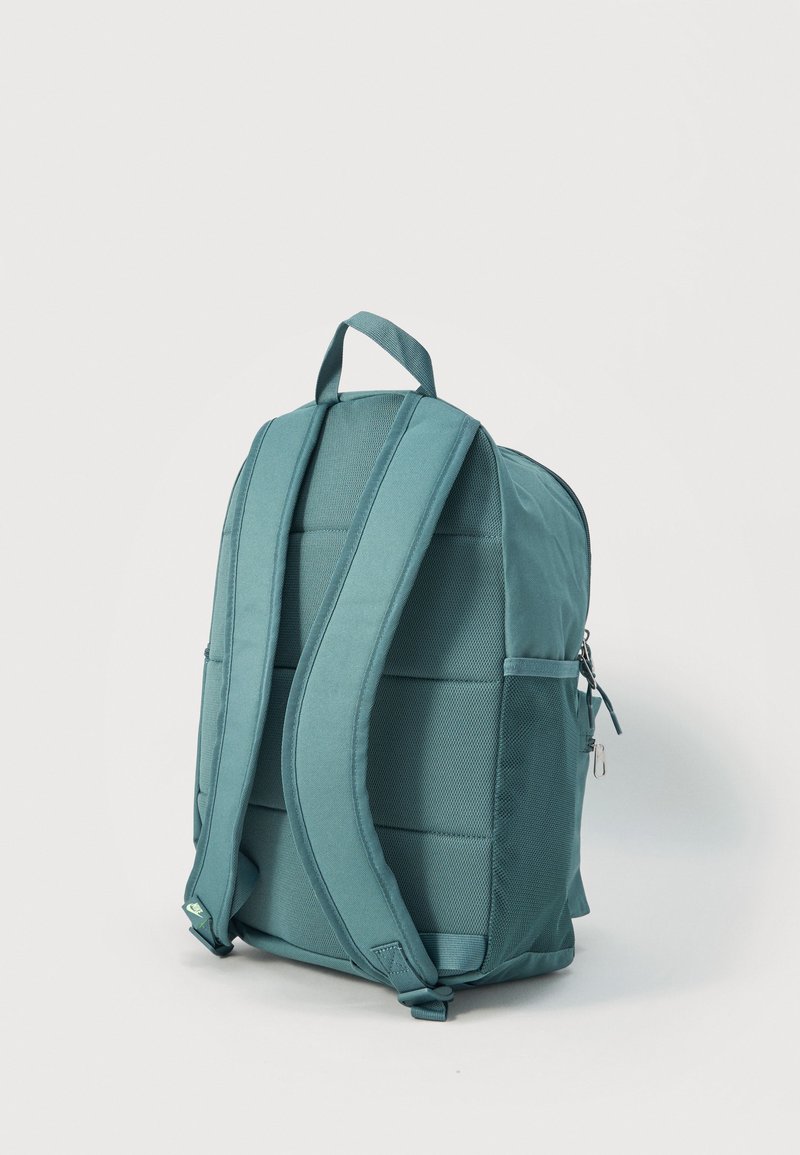 Mochila verde azulado con correas acolchadas para los hombros, asa superior y bolsillos laterales de malla, de pie contra un fondo claro y liso.