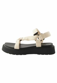 Bež tkanina sandale s crnom gumiranom potplatom, s višestrukim ukriženim trakama i podesivim kopčanjem. Elegantni, minimalistički dizajn.