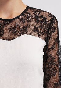 Lace top with black floral pattern on sleeves, contrasting white body, rounded neckline, and textured fabric.