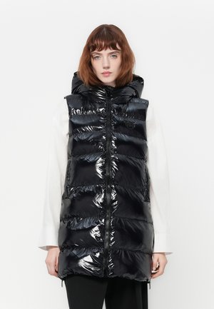 Vrouw in een glanzend zwarte puffervest met capuchon, zip-up over een langemouwen wit shirt, staande tegen een plain witte achtergrond.