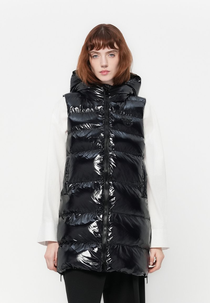 Vrouw in een glanzend zwarte puffervest met capuchon, zip-up over een langemouwen wit shirt, staande tegen een plain witte achtergrond.