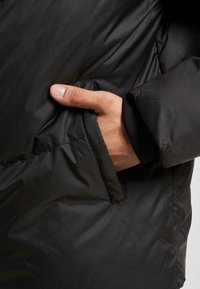 Sort pufferjakke med sidelomme. Materialet er glat og skinnende, med ribbede manchetter ved håndleddet.