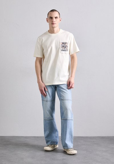 Kreem värvi lühikeste varrukatega T-särk, millel on tasku graafika, koos helesiniste sirgete pükstega ja heleda tekstuuriga klotsidega.