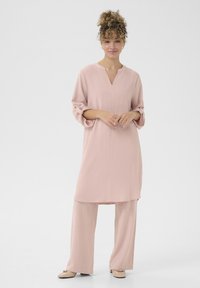 Tunic roz deschis cu decolteu în V și mâneci rulabile, combinată cu pantaloni lejeri asortați. Material neted și fluid, cu o textură subtilă.