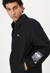 Black zip-up jacket featuring a collar, chest pocket, and elastic cuffs. Includes a graphic accent on the sleeve with Lacoste branding.
