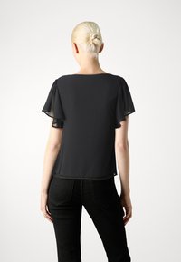 Svart topp med korte ermer og transparente volang-ermer, glatt tekstur, rund halslinning og løs passform, sammen med mørke jeans.