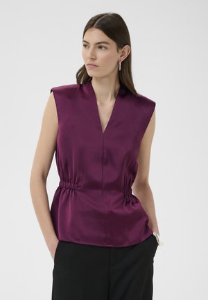 Femme portant un haut sans manches violet profond avec col en V et taille froncée, pantalon noir et boucles d’oreilles argentées en forme de créoles, debout les mains dans les poches.