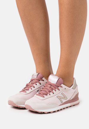 Piedi che indossano sneakers New Balance rosa chiaro e malva con dettagli in mesh e suede su sfondo bianco.