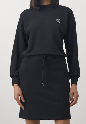 Femme portant une robe noire à manches longues avec un petit logo blanc brodé sur la poitrine et une taille à cordon.