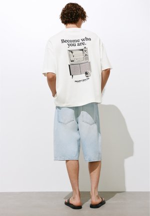 Persoană cu păr creț poartă un tricou alb oversized cu textul „Fii cine ești” și imprimeu de televizor vintage, pantaloni scurți de denim deschis la culoare și sandale negre.
