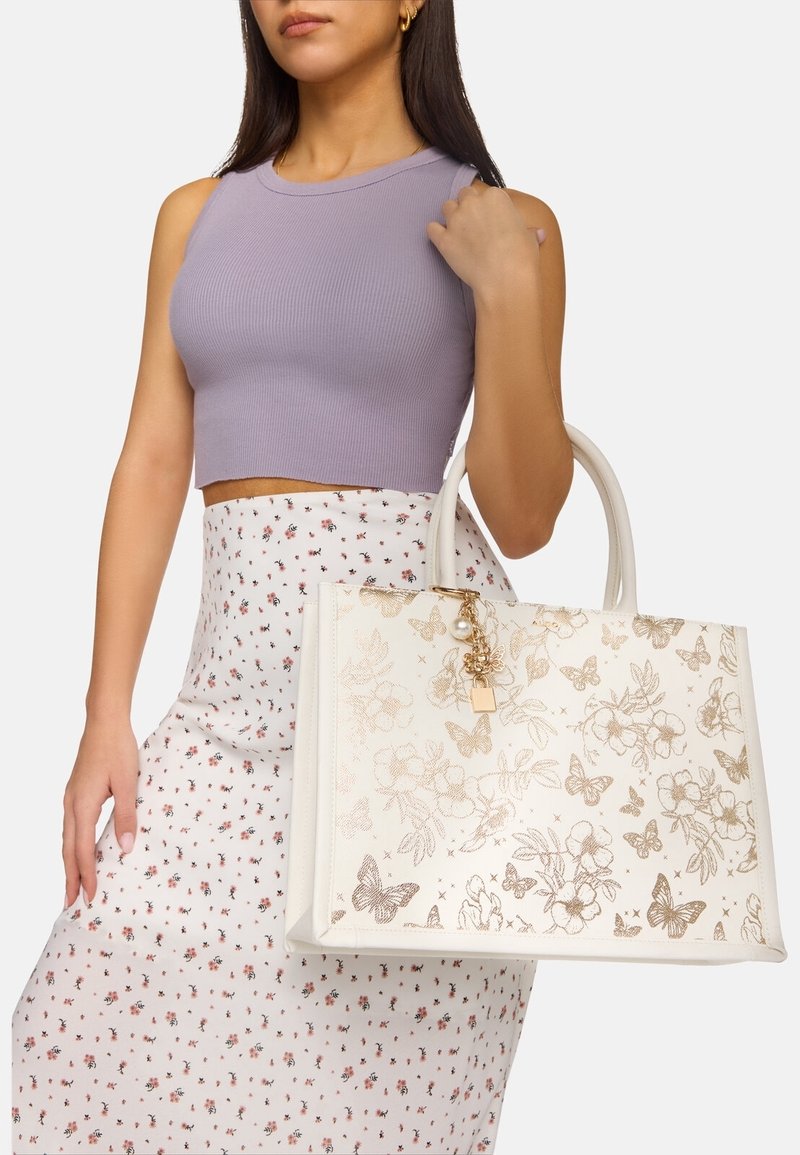 Femme portant un haut sans manches lavande et une jupe blanche fleurie tenant un sac à main blanc avec des motifs dorés de papillons et de fleurs.