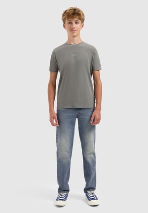 Tienerjongen staat, draagt een grijs T-shirt met kleine witte tekst, vervaagde blauwe jeans en blauwe sneakers met witte veters, tegen een effen achtergrond.