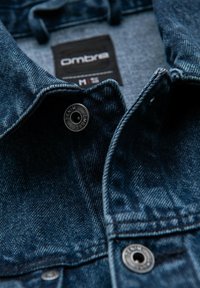 Giacca di jeans con una tonalità blu scuro, chiusura con bottoni e tessuto testurizzato. L'etichetta riporta "Ombre" con l'indicazione della taglia. Design classico del colletto.