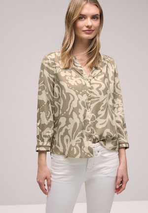 Jeune femme aux cheveux blonds et raides portant un chemisier boutonné à motifs abstraits beige et crème ainsi qu'un pantalon blanc, sur un fond uni.