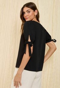 Blouse noire à manches courtes avec des manches bouffantes, attaches latérales aux poignets, texture lisse et encolure arrondie. Vue arrière affichée.