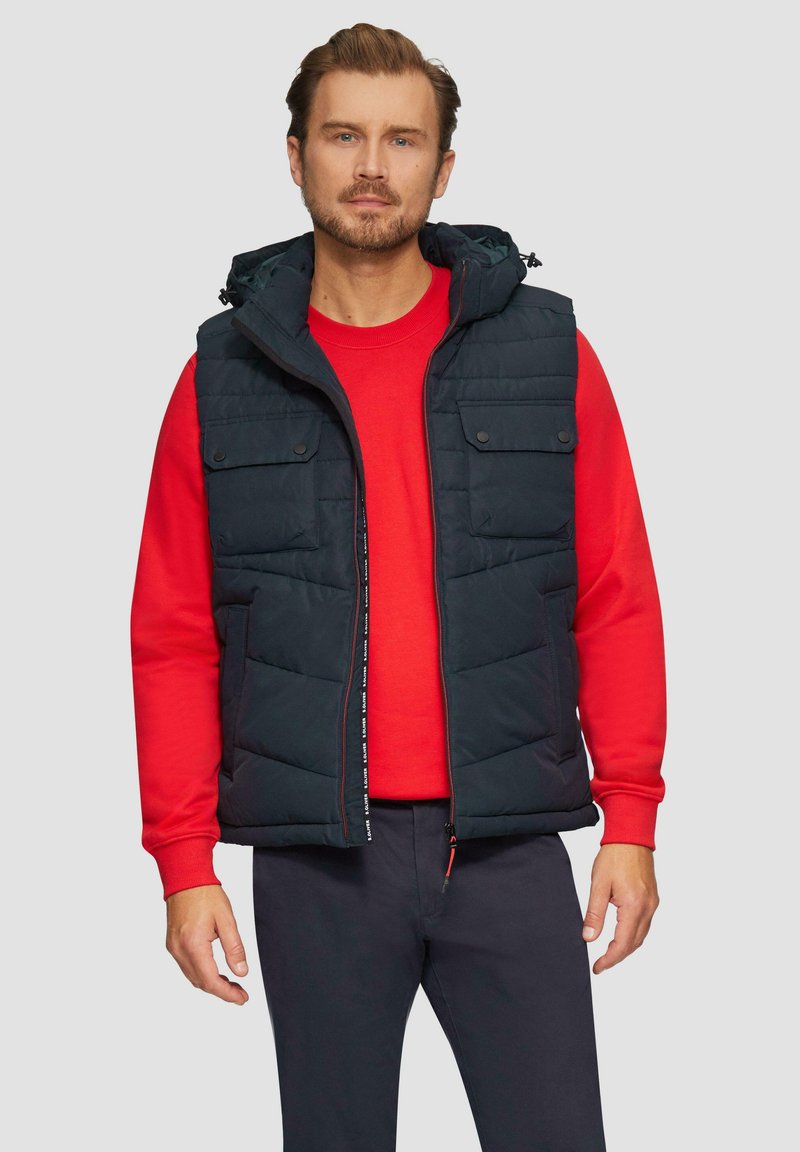 Marineblaue Steppweste mit Kapuze, Reißverschluss und Fronttaschen, über einem roten Sweatshirt getragen, kombiniert mit dunklen Hosen.