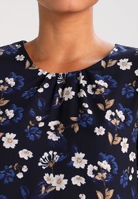 Blusa blu navy con motivo floreale bianco e blu, con scollatura arricciata e maniche corte. Tessuto liscio con una consistenza morbida.