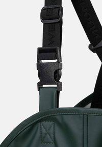 Gros plan sur une boucle en plastique noir attachée à une sangle noire et à un sac vert, montrant le mécanisme de fermeture et le détail du tissu cousu.