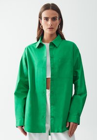 Vihreä nappikauluksellinen paita, jossa on pitkät hihat, vasemmassa rinnassa yksi tasku ja rento leikkaus. Sileä, kevyt kangas.