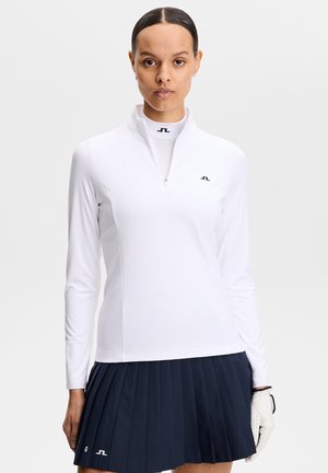 Donna che indossa una maglia bianca a maniche lunghe con zip e una gonna plissettata blu navy, con un guanto da golf bianco sulla mano sinistra.