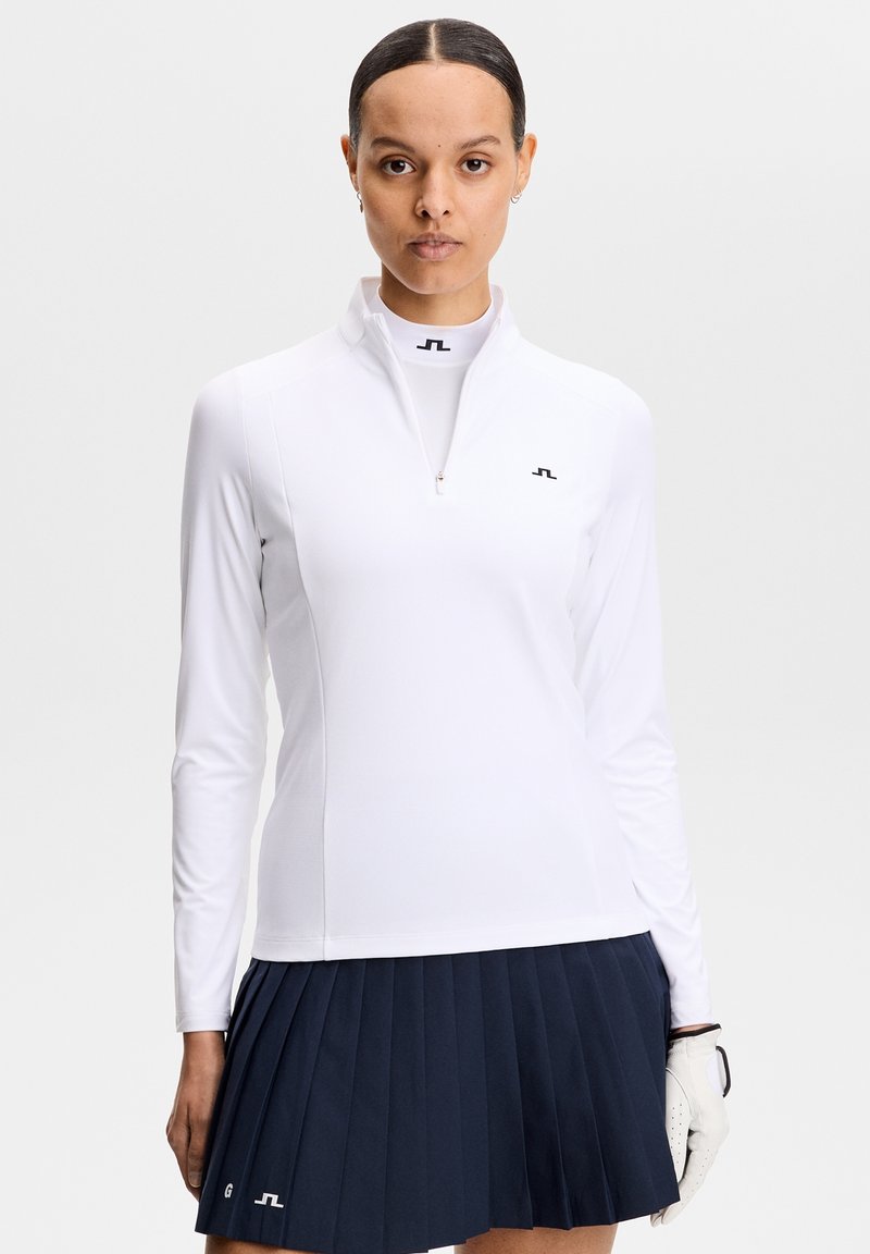 Vrouw met een wit ritsvest met lange mouwen en een marineblauwe geplooide rok, met een witte golfhandschoen aan haar linkerhand.