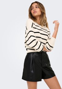Femme en pull crème avec des rayures noires et un short en cuir noir, posant avec un bras plié et regardant sur le côté.