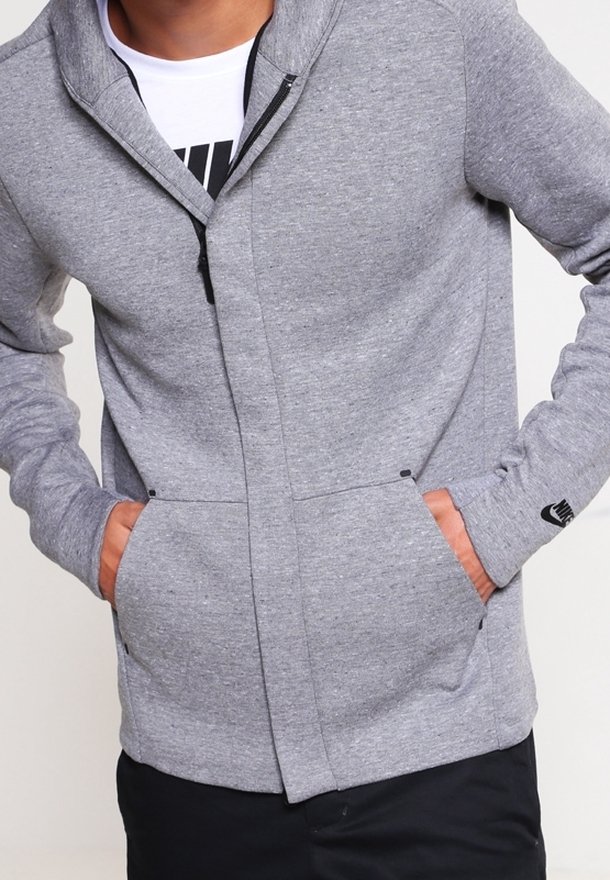 Homme portant un sweat à capuche zippé gris clair avec les mains dans les poches avant, un logo Nike noir sur le poignet de la manche gauche et un t-shirt blanc en dessous.