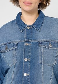 Femme portant une veste en jean bleue décolorée avec des boutons en métal et des poches à rabat sur la poitrine, montrée des épaules jusqu'au milieu du torse.