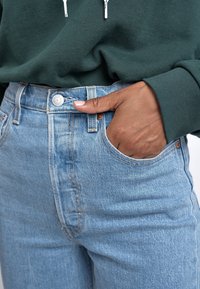 Vaqueros de denim azul claro de cintura alta, con cierre de botones y bolsillo delantero. Acompañados de una sudadera con capucha en verde oscuro.