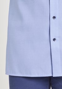 Camicia blu chiaro a piccoli quadretti con bottoni blu scuro indossata sopra pantaloni blu scuro, primo piano sulla parte inferiore del torso.