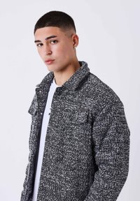 Giovane uomo con i capelli corti e scuri indossa una giacca a trama bianca e nera sopra una camicia bianca, guardando leggermente di lato su uno sfondo semplice.