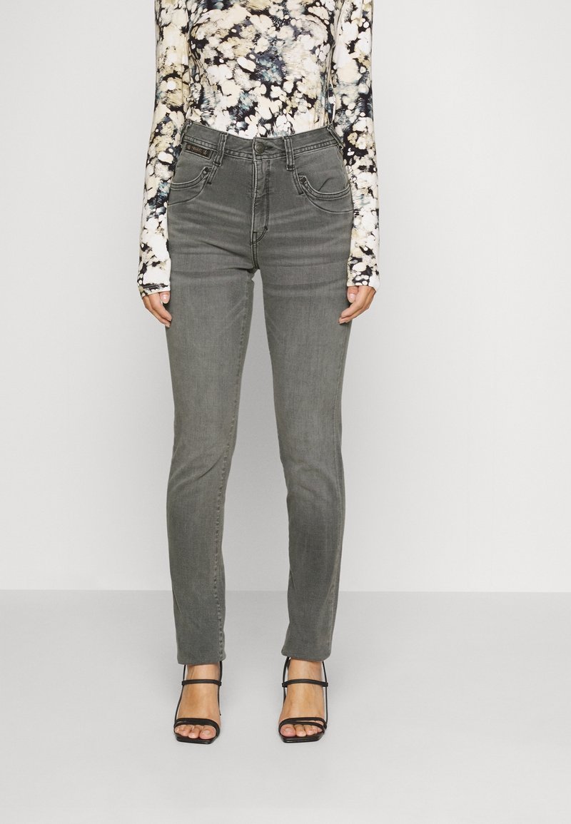 Femme portant un jean skinny gris taille haute, des talons noirs à brides et un haut ajusté à manches longues à motif floral sur un fond clair uni.