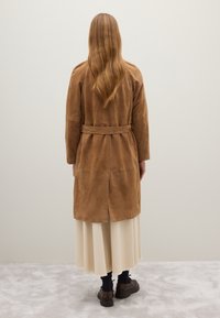 Cappotto in suede marrone, lungo fino al ginocchio, con vita alta cinturata, dotato di uno spacco sul retro e una superficie liscia, indossato sopra una gonna crema e scarpe marroni.