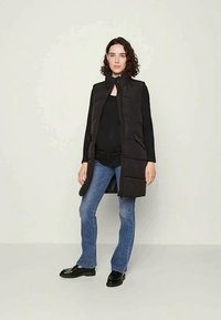 Svart puffervest med høy krage, glidelåslukking og frontlommer. Kombinert med en svart topp og blå jeans. Flate sko fullfører antrekket.