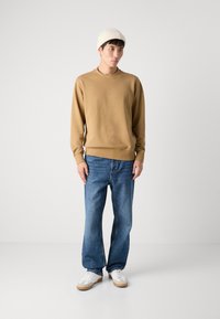 Sudadera de algodón marrón con puños acanalados, combinada con jeans de mezclilla azules y zapatillas blancas con suelas beige. Gorro de punto color crema.