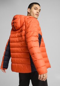 Polstret jakke i lys orange med sorte detaljer; har hætte og quiltet design. Stoffet ser glat og let reflekterende ud.