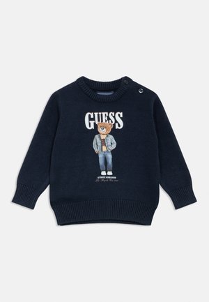 Pulover bleumarin cu manșete și tiv striate, cu un grafic cu urs în denim și textul "GUESS" pe partea din față. Detaliu cu nasturi pe umăr.