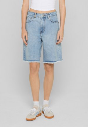 Person iført lyseblå denim shorts til knæet med frynset kant, hvid kort top, hvide sokker og beige sneakers.