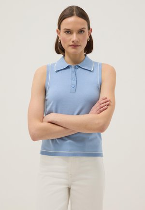 Femme aux cheveux bruns courts portant des boucles d'oreilles anneaux dorées, un haut bleu clair sans manches à col, et un pantalon blanc, bras croisés, fond neutre.