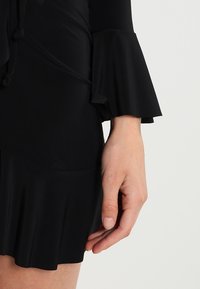 Czarna sukienka z rozkloszowanymi rękawami, o gładkiej fakturze i falbaniastym dołem, prezentująca prosty i elegancki krój.