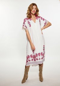 Weißes Kleid mit rotem Blumenstickerei und Quasten an den Ärmeln. Verfügt über einen V-Ausschnitt und ein lockeres Design, kombiniert mit braunen Stiefeletten.