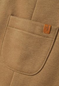 Brūna auduma ar teksturētu virsmu, kurā ir liela kabata un nelaba ādas etiķete, uz kuras ir iepresēts "Lilt Atelier".