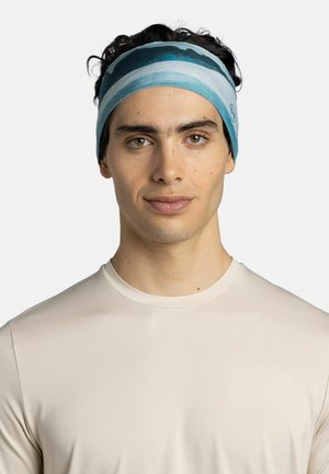 Young man with curly dark hair wearing a blue striped headband and a plain beige long-sleeve shirt, facing forward with a neutral expression.