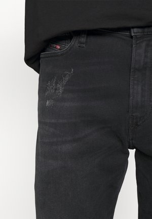 Gros plan d'une personne portant un jean noir déchiré avec un petit détail d'usure estompée près de la poche avant et une chemise noire rentrée.