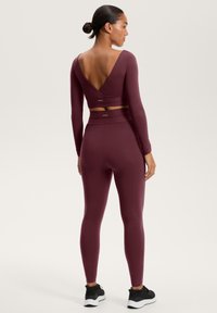 Set de îmbrăcăminte sport în nuanță de vișiniu, compus dintr-o bluză cu mânecă lungă și un decolteu în V adânc la spate și leggings asortați. Textură netedă cu detalii de branding minime.