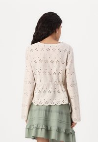 Light beige long-sleeve top with floral cut-out patterns, gathered waist, and scalloped hem, paired with a layered green ruffled skirt.