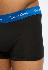 Κοντινό πλάνο ενός ατόμου που φοράει μαύρα ανδρικά εσώρουχα boxer με μπλε ζώνη μέσης που φέρει το εμπορικό σήμα Calvin Klein με λευκό κείμενο.