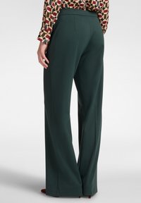 Vrouw in donkergroene wijde broek en een gestreept shirt met lange mouwen, staand tegen een witte achtergrond.